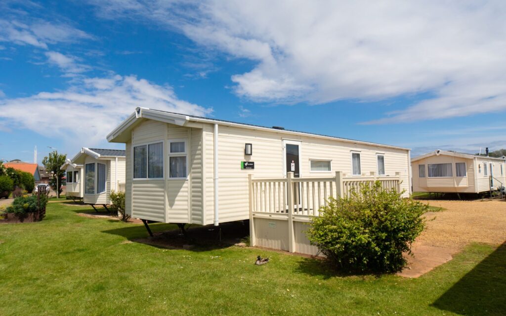 Cream coloured modern static caravan with a veranda and bright blue sky sprinkled with clouds.