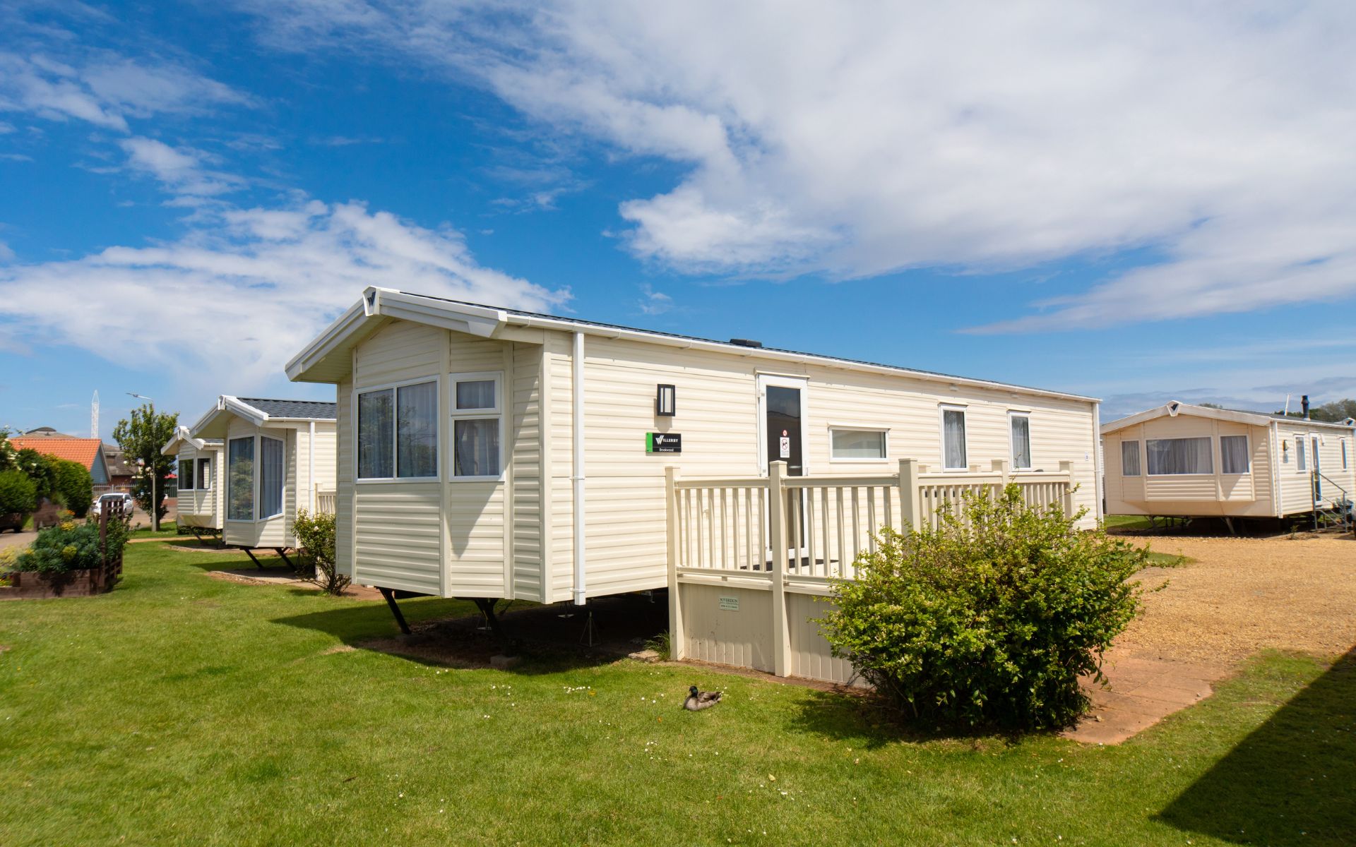 Cream coloured modern static caravan with a veranda and bright blue sky sprinkled with clouds.