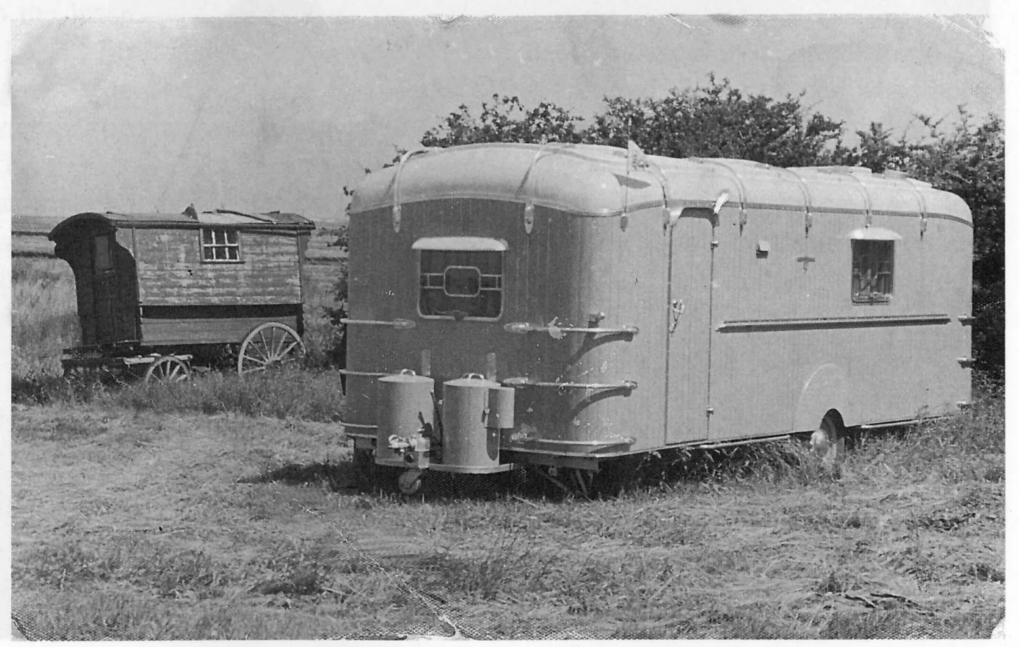Black and white image of an early metal caravan from around the 1950's with an original wooden horse drawn caravan in the background.