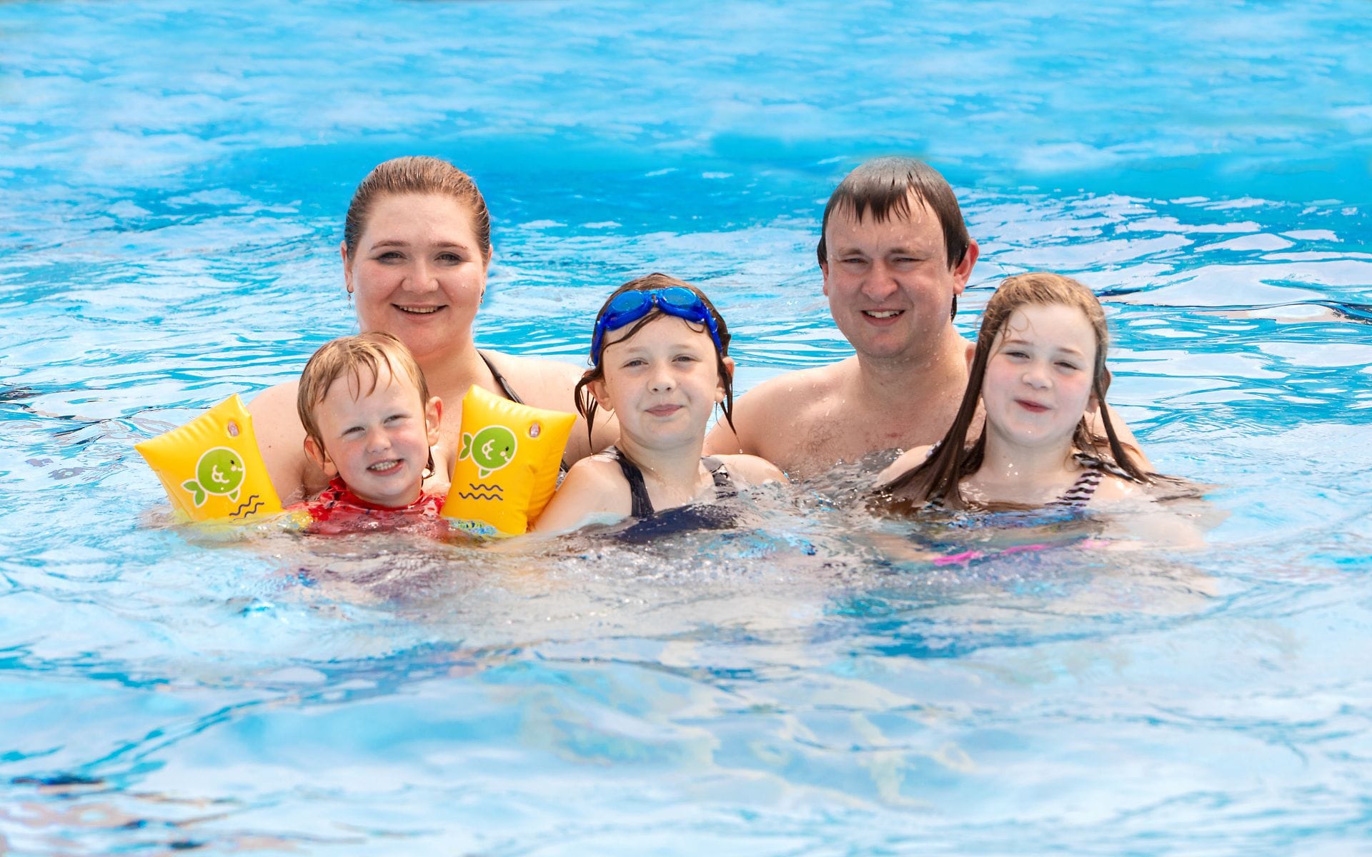 A family of five enjoys a pool day. Parents and three children smile in the bright blue water. The mood is joyful and relaxed.