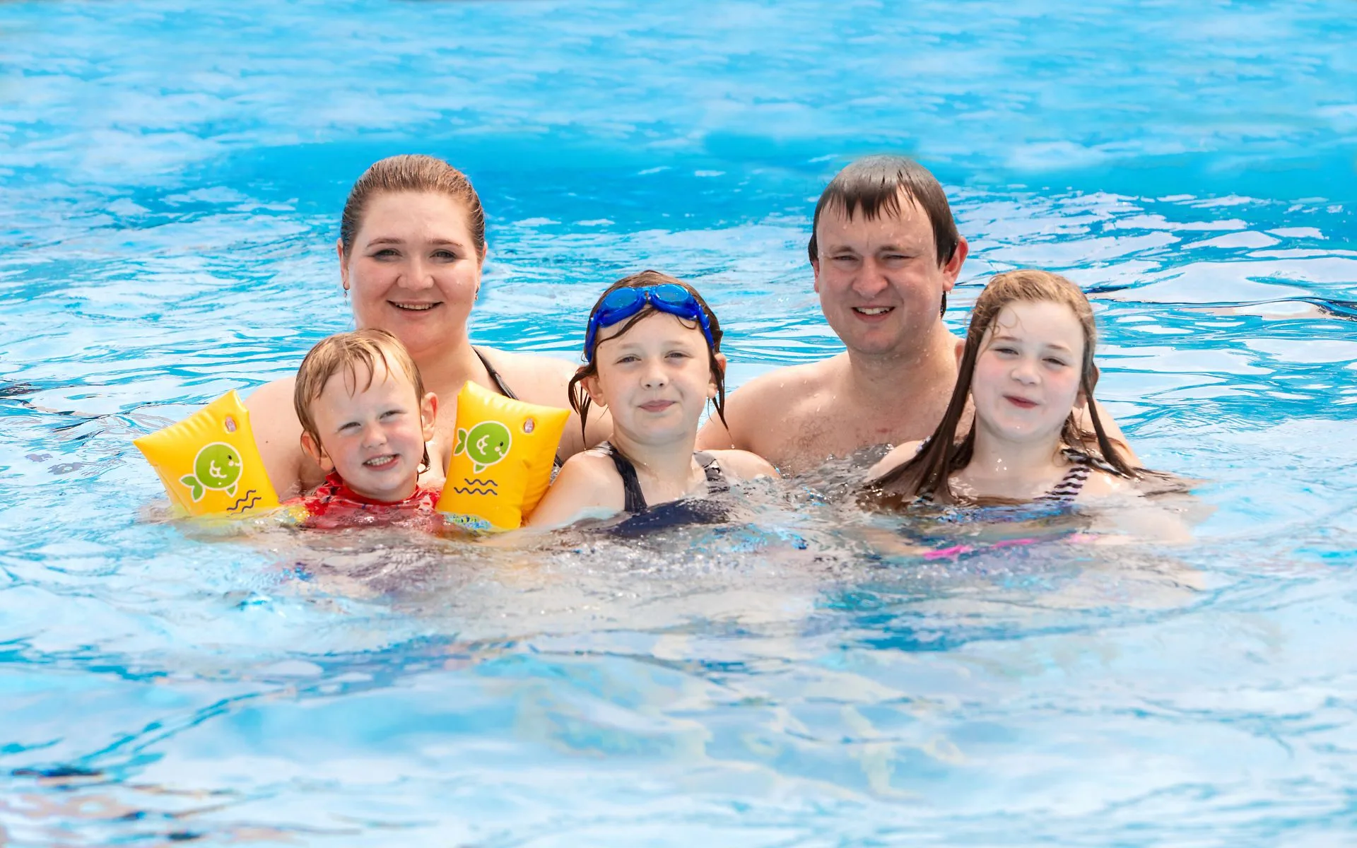A family of five enjoys a pool day. Parents and three children smile in the bright blue water. The mood is joyful and relaxed.